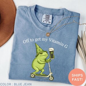 Op de afbeelding: Blauw spijkerbroek Comfort Colors T-shirt met de tekst "Off to get my Vitamin G". De afbeelding toont een groene kikker met een hoed en bier, op een scooter. Een gouden ketting ligt op het T-shirt.