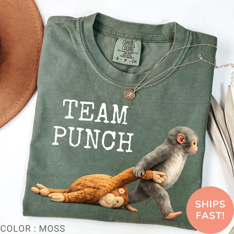 May include: Moss green Comfort Colors t-shirt with the text "TEAM PUNCH" in white. A cartoon monkey is pulling another monkey. A silver necklace is draped across the shirt. The shirt is labeled "S - P - CH".