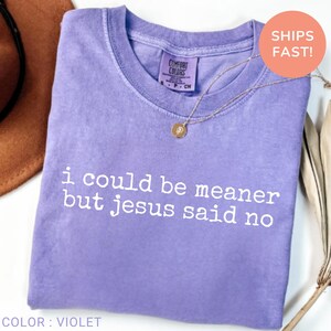 May include: A violet-colored t-shirt with the text "i could be meaner but jesus said no" in white. The shirt is folded and has a gold necklace draped over it. The shirt is labeled "Comfort Colors".