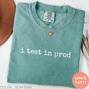 May include: Seafoam green t-shirt with the text "i test in prod" in white. The shirt has a crew neck and short sleeves. A silver necklace with a pendant is draped on the shirt. The shirt is labeled "Comfort Colors".