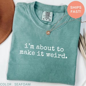 May include: A seafoam green short-sleeve t-shirt with a crew neckline and the white text "i'm about to make it weird." printed on the front. The shirt features a Comfort Colors brand label at the inner neck and is paired with a silver-toned necklace.
