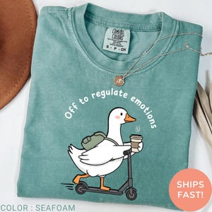 May include: Seafoam green t-shirt with a cartoon goose riding a scooter, holding a coffee cup, and wearing a backpack. The text "Off to regulate emotions" is above the goose. The shirt has a Comfort Colors tag.
