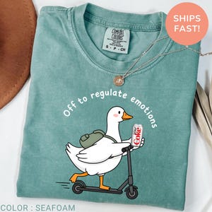 May include: Seafoam green t-shirt featuring a white goose on a scooter, holding a Diet Coke can, with the text "Off to regulate emotions". The shirt has a Comfort Colors label. The colour is described as seafoam.