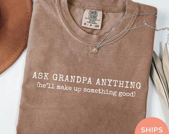 May include: Espresso-colored t-shirt with the text "ASK GRANDPA ANYTHING (he'll make up something good)" in white. The shirt has a crew neck and a small tag near the collar. A silver necklace with a pendant is draped across the shirt.