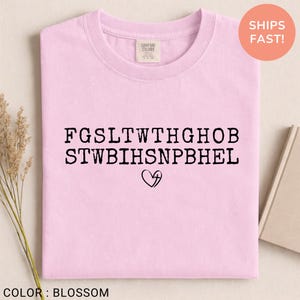 Pu&ograve; includere: Una maglietta rosa chiaro con testo nero e un simbolo a forma di cuore. Il testo &egrave; disposto su due righe di lettere maiuscole. La maglietta ha uno scollo tondo e un'etichetta "Comfort Colors". Il colore della maglietta &egrave; "Blossom".