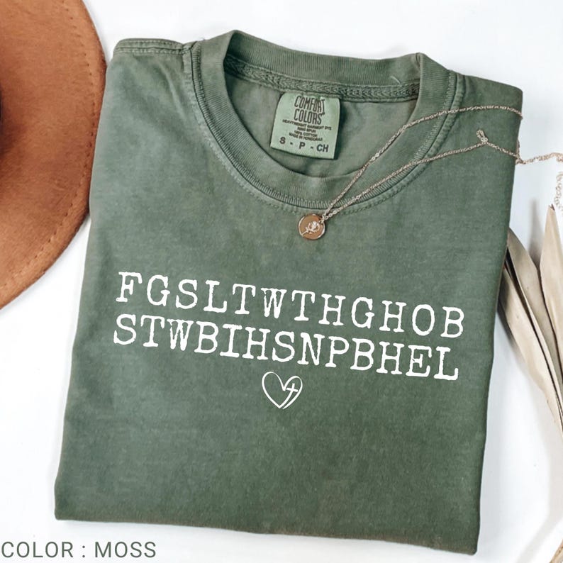Pu&ograve; includere: Una maglietta Comfort Colors verde muschio con testo bianco e un simbolo a forma di cuore. Una collana d'oro con un ciondolo &egrave; drappeggiata sulla maglietta. La maglietta &egrave; piegata e appoggiata su una superficie bianca.