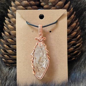 May include: A clear, pointed quartz crystal pendant wrapped in copper wire, suspended from a black cord. The pendant is mounted on a rectangular brown cardboard display card. The wire wrapping features intricate coils and a decorative knot design.