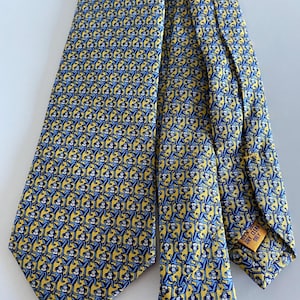 Puede incluir: Una corbata amarilla con un patrón repetido de monos azules. La corbata tiene una forma clásica y está hecha de una tela suave. La etiqueta dice "Limpiar en seco solamente".
