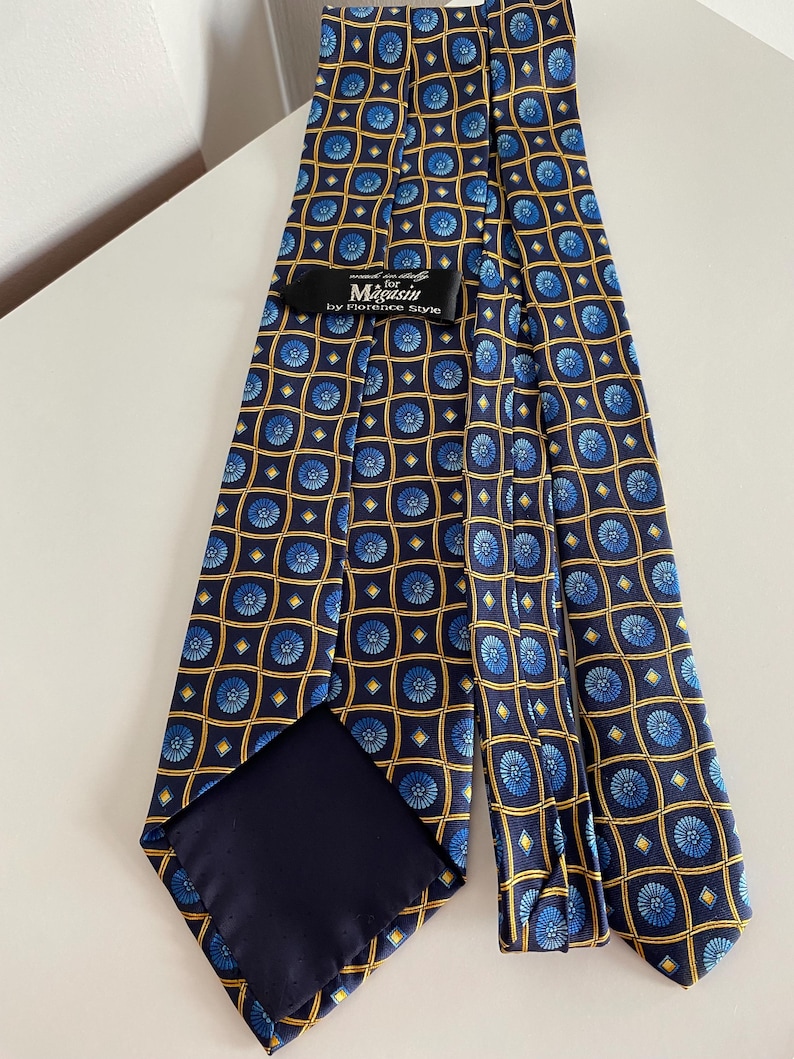Puede incluir: Una corbata azul marino con un patr&oacute;n repetido de dise&ntilde;os florales azules dentro de cuadrados dorados. La etiqueta dice "Magasin by Florence Style."