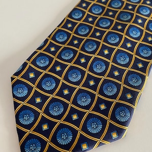 Puede incluir: Corbata azul marino con un patr&oacute;n repetido de dise&ntilde;os florales azules y formas geom&eacute;tricas doradas. La corbata est&aacute; hecha de una tela suave y es adecuada para vestimenta formal o de negocios.