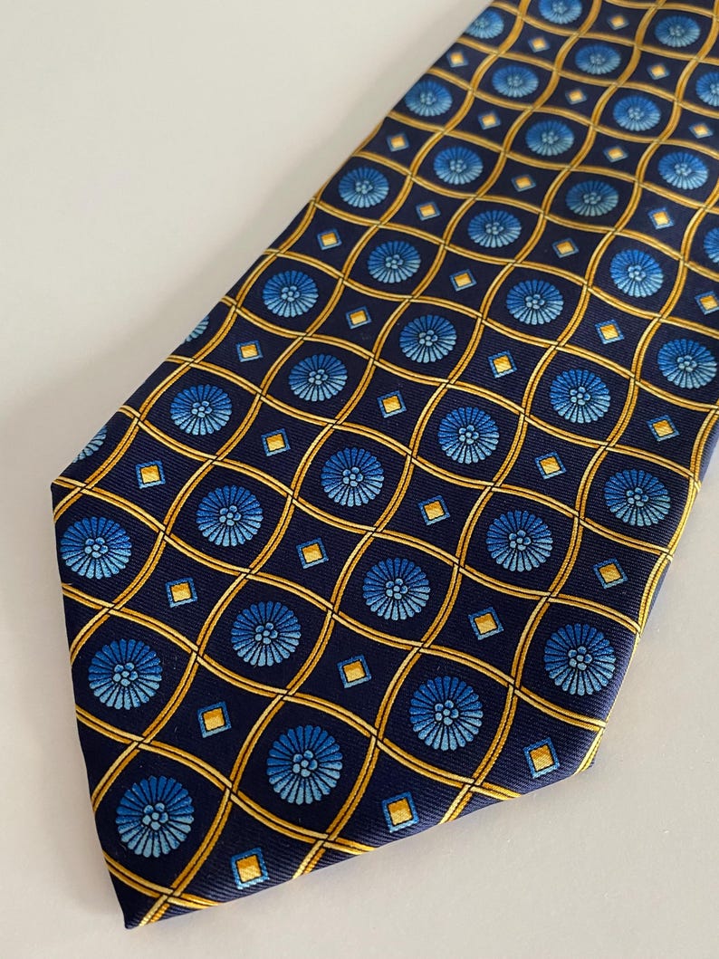 Puede incluir: Corbata azul marino con un patr&oacute;n repetido de dise&ntilde;os florales azules y formas geom&eacute;tricas doradas. La corbata est&aacute; hecha de una tela suave y es adecuada para vestimenta formal o de negocios.