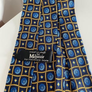 Puede incluir: Una corbata azul marino con un patr&oacute;n repetido de dise&ntilde;os florales azules dentro de cuadrados y diamantes dorados. La etiqueta dice "made in Italy for Magasin by Florence Style."