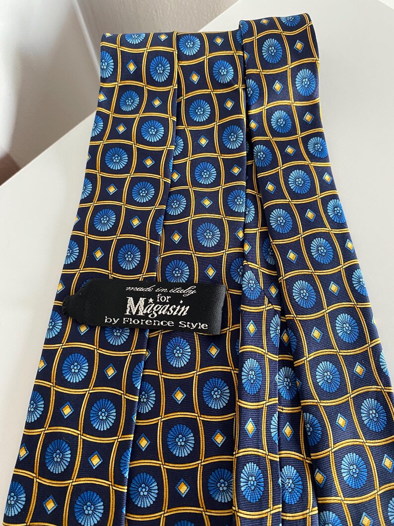 Puede incluir: Una corbata azul marino con un patr&oacute;n repetido de dise&ntilde;os florales azules dentro de cuadrados y diamantes dorados. La etiqueta dice "made in Italy for Magasin by Florence Style."
