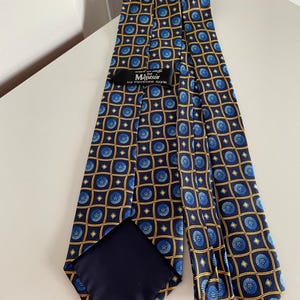 Puede incluir: Una corbata azul marino con un patr&oacute;n repetitivo de dise&ntilde;os florales azules dentro de cuadrados dorados. La corbata tiene un reverso azul oscuro y una etiqueta que dice "Magasin by Florence Style."
