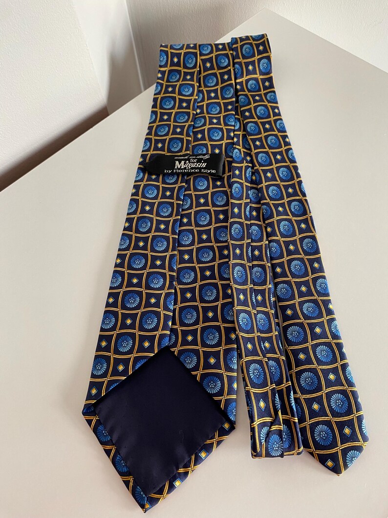Puede incluir: Una corbata azul marino con un patr&oacute;n repetitivo de dise&ntilde;os florales azules dentro de cuadrados dorados. La corbata tiene un reverso azul oscuro y una etiqueta que dice "Magasin by Florence Style."