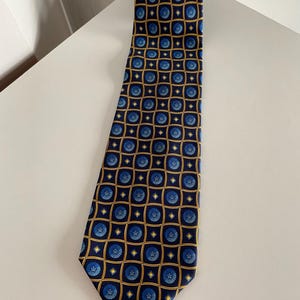Puede incluir: Una corbata azul marino con un patr&oacute;n repetido de dise&ntilde;os florales azules dentro de cuadrados dorados. La corbata est&aacute; hecha de una tela suave y se muestra sobre una superficie blanca. Un accesorio cl&aacute;sico.
