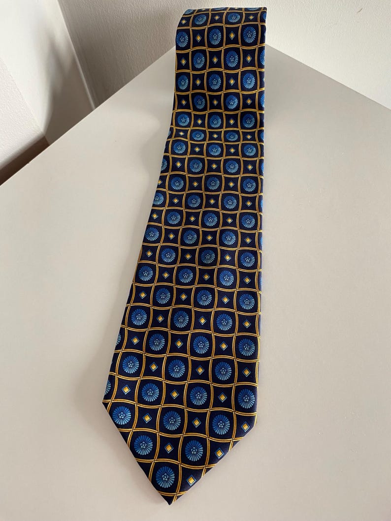 Puede incluir: Una corbata azul marino con un patr&oacute;n repetido de dise&ntilde;os florales azules dentro de cuadrados dorados. La corbata est&aacute; hecha de una tela suave y se muestra sobre una superficie blanca. Un accesorio cl&aacute;sico.