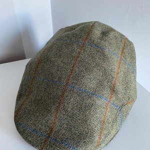 Puede incluir: Gorra plana de tweed verde con un patrón de cuadros de rayas naranjas y azules. La gorra tiene una corona redondeada y una visera corta y rígida. La tela tiene un aspecto texturizado.