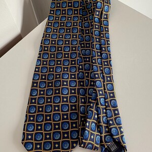 Puede incluir: Una corbata azul marino con un patr&oacute;n repetido de dise&ntilde;os florales azules dentro de cuadrados dorados. La corbata est&aacute; hecha de una tela suave y est&aacute; doblada sobre una superficie blanca.