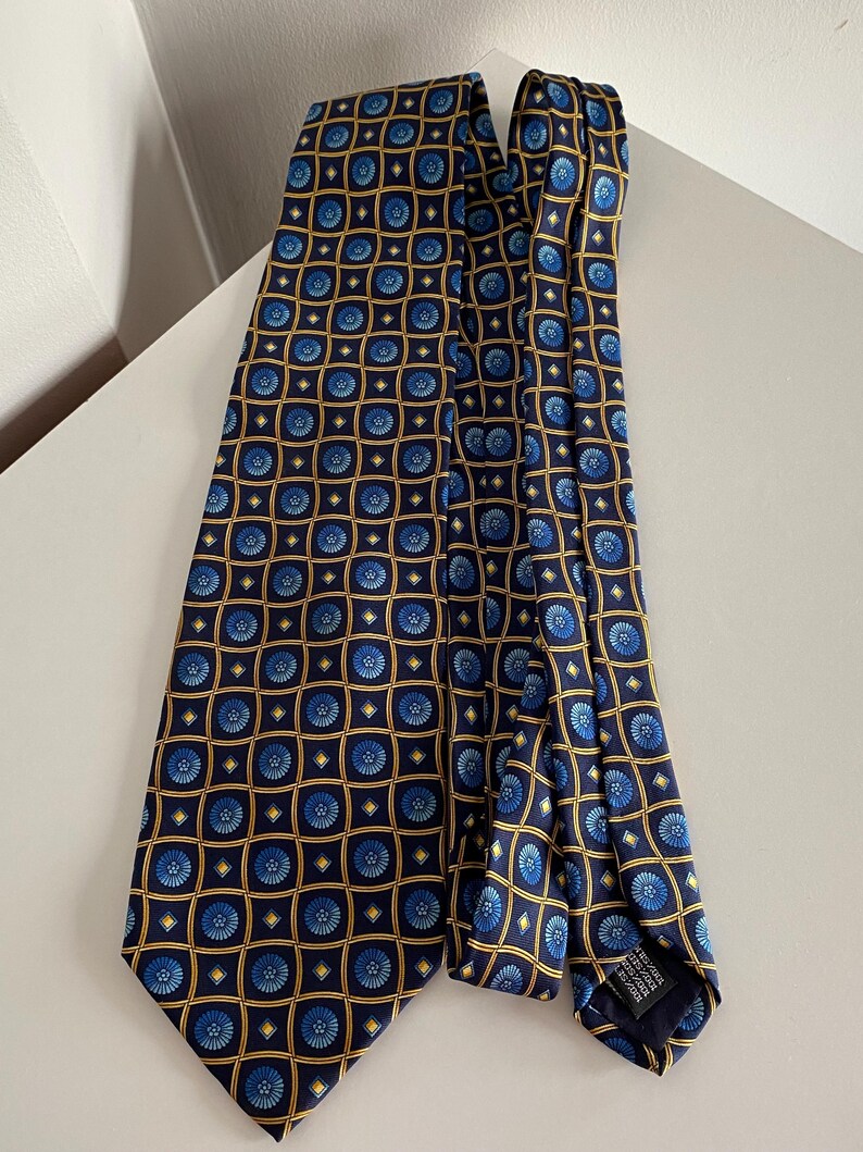 Puede incluir: Una corbata azul marino con un patr&oacute;n repetido de dise&ntilde;os florales azules dentro de cuadrados dorados. La corbata est&aacute; hecha de una tela suave y est&aacute; doblada sobre una superficie blanca.