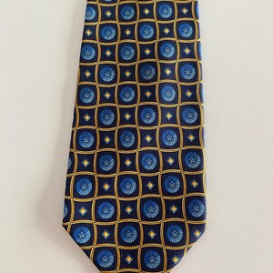 Puede incluir: Una corbata azul marino con un patr&oacute;n repetido de motivos florales azules dentro de cuadrados y diamantes dorados. La corbata, probablemente de seda, es adecuada para ocasiones formales o de negocios. El dise&ntilde;o es sim&eacute;trico.