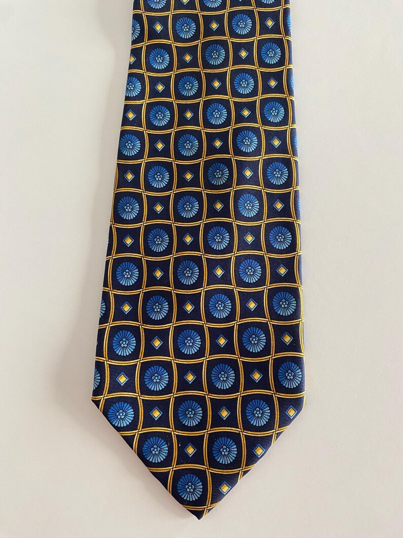 Puede incluir: Una corbata azul marino con un patr&oacute;n repetido de motivos florales azules dentro de cuadrados y diamantes dorados. La corbata, probablemente de seda, es adecuada para ocasiones formales o de negocios. El dise&ntilde;o es sim&eacute;trico.