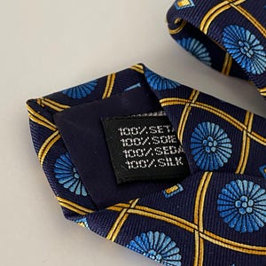 Puede incluir: Corbata de seda azul marino con un patr&oacute;n repetido de dise&ntilde;os florales azules y l&iacute;neas geom&eacute;tricas doradas. La corbata tiene una etiqueta con texto. La tela es 100% seda.