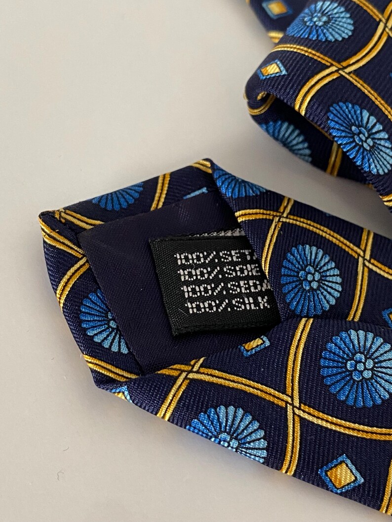 Puede incluir: Corbata de seda azul marino con un patr&oacute;n repetido de dise&ntilde;os florales azules y l&iacute;neas geom&eacute;tricas doradas. La corbata tiene una etiqueta con texto. La tela es 100% seda.