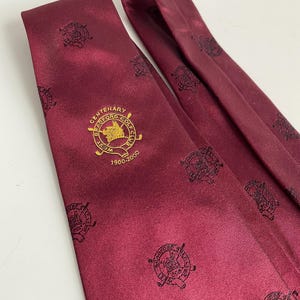 Puede incluir: Corbata burdeos con un patrón repetido de emblemas de club de golf y texto dorado "CENTENARY BRADFORD GOLF CLUB 1900-2000". La corbata está hecha de un material liso, posiblemente seda.