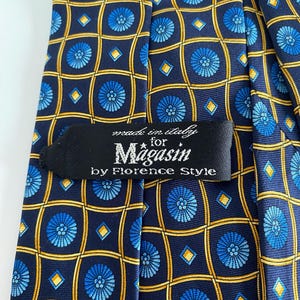 Puede incluir: Corbata de seda azul marino con un patr&oacute;n repetido de medallones florales azules y l&iacute;neas de cuadr&iacute;cula amarillas. La etiqueta dice "made in italy for Magasin by Florence Style."
