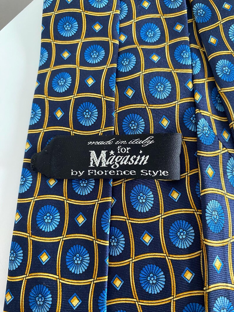Puede incluir: Corbata de seda azul marino con un patr&oacute;n repetido de medallones florales azules y l&iacute;neas de cuadr&iacute;cula amarillas. La etiqueta dice "made in italy for Magasin by Florence Style."