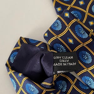 Puede incluir: Corbata de seda azul marino con un patr&oacute;n repetido de dise&ntilde;os florales azules y formas geom&eacute;tricas doradas. La etiqueta dice "DRY CLEAN ONLY MADE IN ITALY".