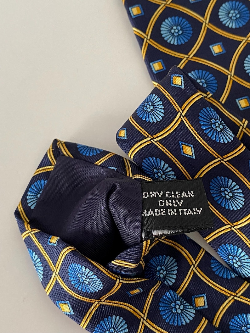Puede incluir: Corbata de seda azul marino con un patr&oacute;n repetido de dise&ntilde;os florales azules y formas geom&eacute;tricas doradas. La etiqueta dice "DRY CLEAN ONLY MADE IN ITALY".