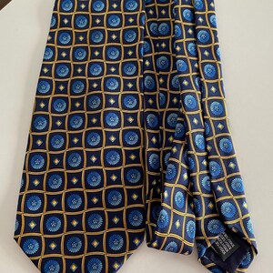 Puede incluir: Corbata de seda azul marino con un patr&oacute;n repetido de dise&ntilde;os florales azules dentro de cuadrados dorados y azul marino. La corbata est&aacute; doblada. Hecha de 100% seda. La corbata est&aacute; sobre una superficie blanca.