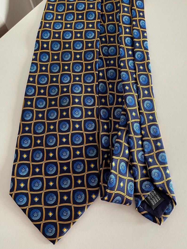 Puede incluir: Corbata de seda azul marino con un patr&oacute;n repetido de dise&ntilde;os florales azules dentro de cuadrados dorados y azul marino. La corbata est&aacute; doblada. Hecha de 100% seda. La corbata est&aacute; sobre una superficie blanca.