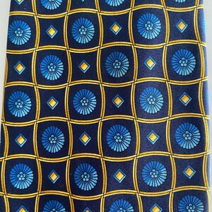 Puede incluir: Corbata azul marino con un patr&oacute;n repetido de motivos florales azules dentro de cuadrados dorados y peque&ntilde;os rombos dorados. La corbata est&aacute; hecha de una tela lisa, posiblemente seda.
