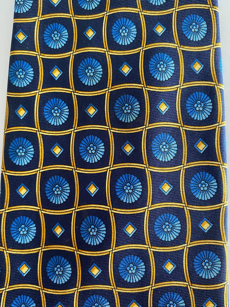 Puede incluir: Corbata azul marino con un patr&oacute;n repetido de motivos florales azules dentro de cuadrados dorados y peque&ntilde;os rombos dorados. La corbata est&aacute; hecha de una tela lisa, posiblemente seda.