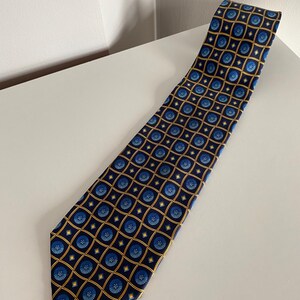 Puede incluir: Corbata azul marino con un patr&oacute;n repetido de dise&ntilde;os florales azules dentro de cuadrados dorados. La corbata est&aacute; hecha de una tela suave y se muestra sobre una superficie blanca. Un accesorio cl&aacute;sico.