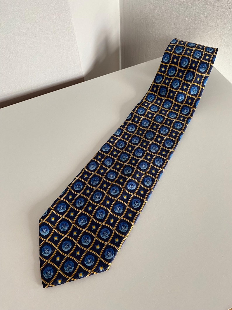 Puede incluir: Corbata azul marino con un patr&oacute;n repetido de dise&ntilde;os florales azules dentro de cuadrados dorados. La corbata est&aacute; hecha de una tela suave y se muestra sobre una superficie blanca. Un accesorio cl&aacute;sico.