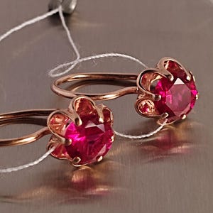 May include: A pair of rose gold earrings with a flower-shaped setting. Each earring features a vibrant, round, red gemstone. The earrings are displayed on a reflective surface, with a white string attached.