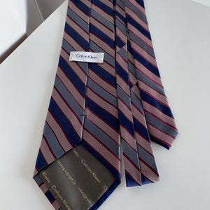 Puede incluir: Una corbata Calvin Klein con un diseño a rayas en gris, azul marino y burdeos. La corbata presenta el logotipo de Calvin Klein en una etiqueta blanca.