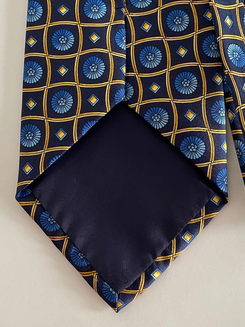 Puede incluir: Una corbata azul marino con un patr&oacute;n repetitivo de dise&ntilde;os florales azules y formas geom&eacute;tricas amarillas. El interior de la corbata es de tela azul marino lisa. Un accesorio cl&aacute;sico.