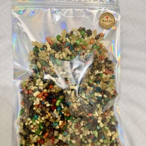 Op de afbeelding: Een doorzichtige plastic zak gevuld met verschillende kleurrijke harsstukjes. De zak heeft een klein rond sticker met de woorden "Bedankt" en een hart.