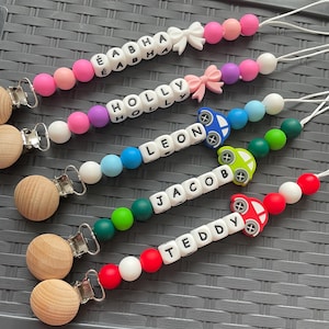 May include: Five personalised dummy clips featuring round silicone beads in various colours, white letter beads spelling the names Eabha, Holly, Leon, Jacob, and Teddy, wooden clips, and decorative silicone shapes including bows and cars.