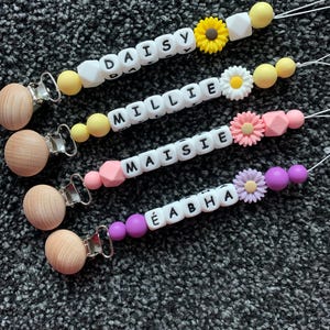 Puede incluir: Cuatro mordedores de bebé de silicona de colores con clips de madera. Cada mordedor tiene un nombre diferente escrito en cuentas blancas y un amuleto de flor. Los nombres son Daisy, Millie, Maisie y Éabha.