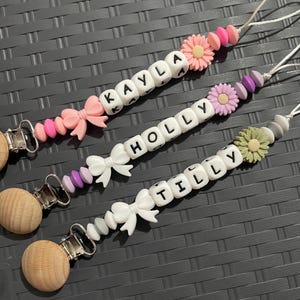 May include: Three personalized pacifier clips with names spelled out in white letter beads. Each clip features a different color scheme, including pink, purple, and green accents, along with decorative bows and flower-shaped beads. The clips have wooden pacifier holders.
