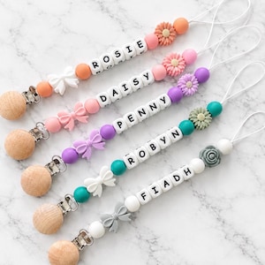 Könnte beinhalten: Fünf personalisierte Schnullerketten mit Holzclips, weißen Buchstabenperlen mit den Namen ROSIE, DAISY, PENNY, ROBYN und FIADH, bunten Silikonperlen sowie dekorativen Silikonformen wie Schleifen und Blumen in Orange, Pink, Lila, Türkis und Grau.
