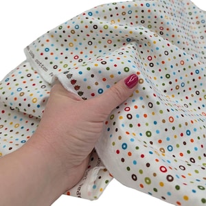May include: A white cotton fabric featuring a repeating pattern of small, multicolored circles in shades of blue, green, orange, and brown. The fabric edge displays a row of small ladybug illustrations and the text 100% cotton Japan.