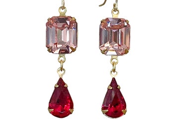 May include: A pair of dangle earrings featuring a rectangular light pink crystal above a teardrop-shaped red crystal. The crystals are set in gold-colored metal and hang from gold-colored ear wires. The earrings are displayed against a white background.