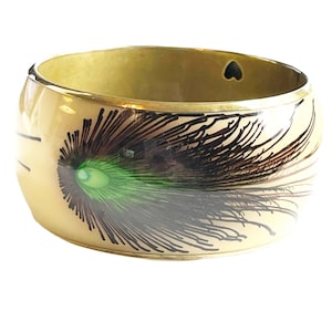 May include: A cream-colored bangle bracelet with a gold-colored metal band and a peacock feather design. The feather is black, brown, and green.